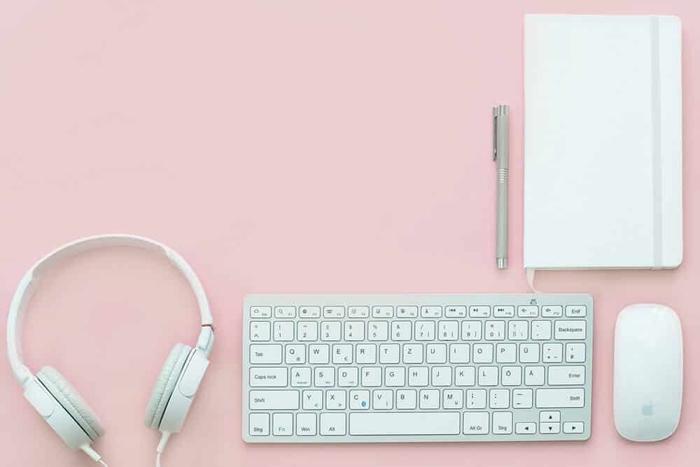 White Headphones, a wireless keyboard and a white journal on a pink writing desk