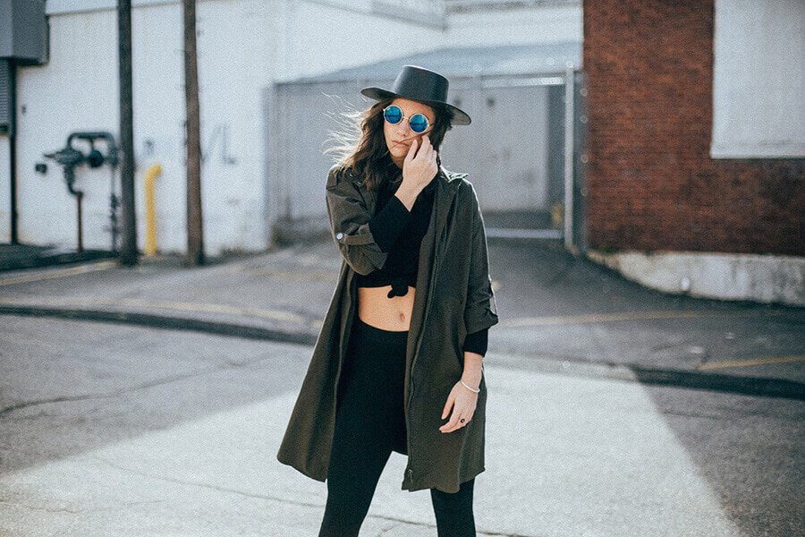 Girl wearing a pair of stretch black pants, a crop top, sunglasses, a hat and a dark green long trench