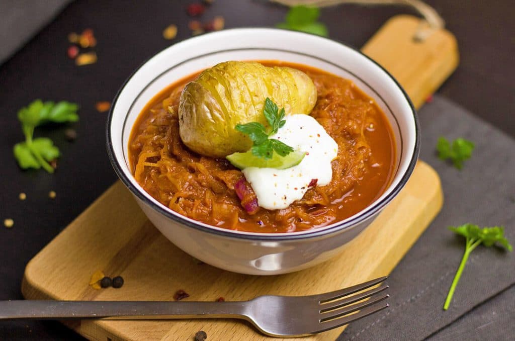 A bowl of bigos with a baked potato, sour cream and cabbage