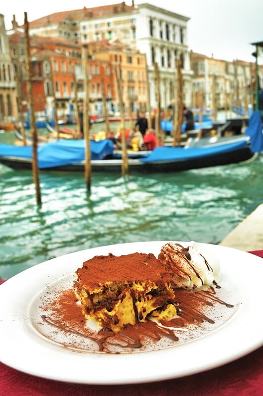 Una porzione di tiramisù servita accanto a un canale a Venezia conle gondole sullo sfondo