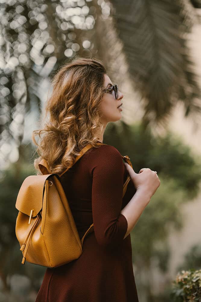 Ragazza bionda con indosso uno zainetto in pelle color senape, occhiali da sole e un cappottino leggero bordeaux