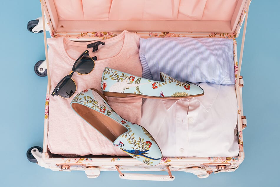 Open suitcase ready for a cruise, with pastel colored shits, stylish shoes and sunglasses