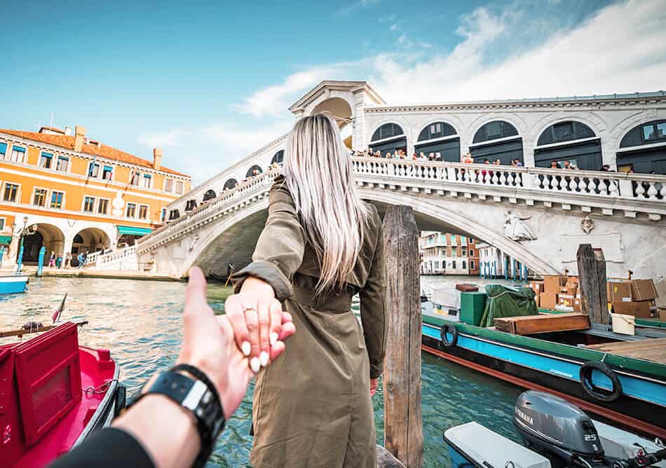 Mergina dėvi tamsiai žalią tranšėją, traukdama savo vaikiną link Rialto tilto Venecijoje (Italija)