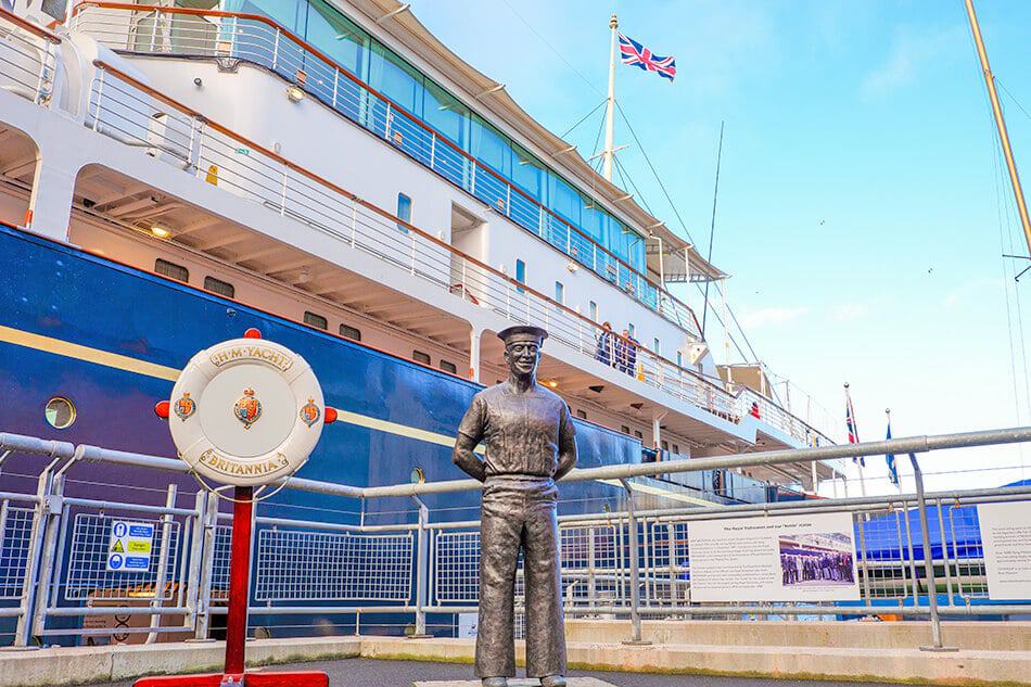 Statua di marinaio accanto al Royal Yacht Britannia e bandiera inglese sullo sfondo