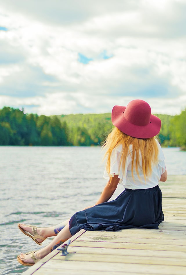 Ragazza con indosso un cappello rosso e gonna lunga seduta sul pontile accanto a un lago di montagna