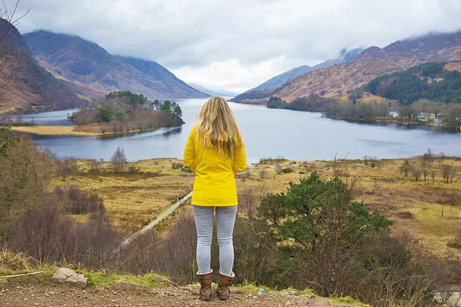 Girl wearing a yellow raincoat standing on a ledge overlooking a lake in Scotland