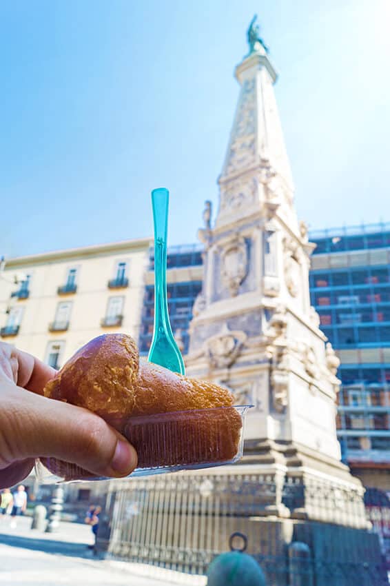 Babà napoletano semplice con forchettina blu davanti a un monumento a Napoli